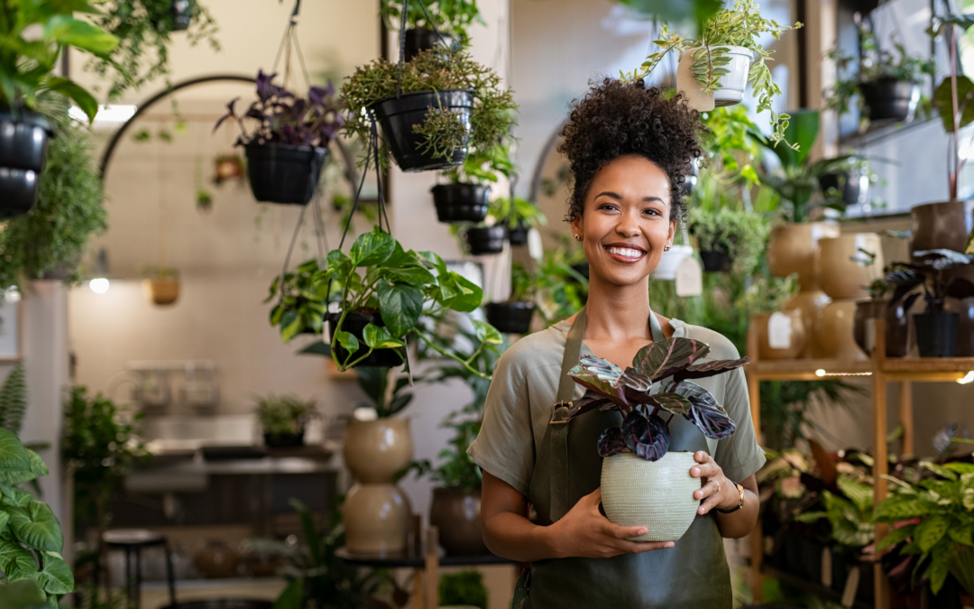 Image shows picture of a woman florist surrounded by plants and greenery, holding a white pot with a plant in it.